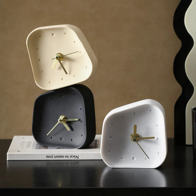 Three minimalist square analog clocks with rounded corners in cream, dark gray, and white, arranged on a black surface with a book and a wavy decorative object in the background.