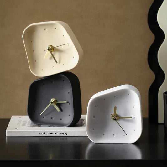Three minimalist square analog clocks with rounded corners in cream, dark gray, and white, arranged on a black surface with a book and a wavy decorative object in the background.